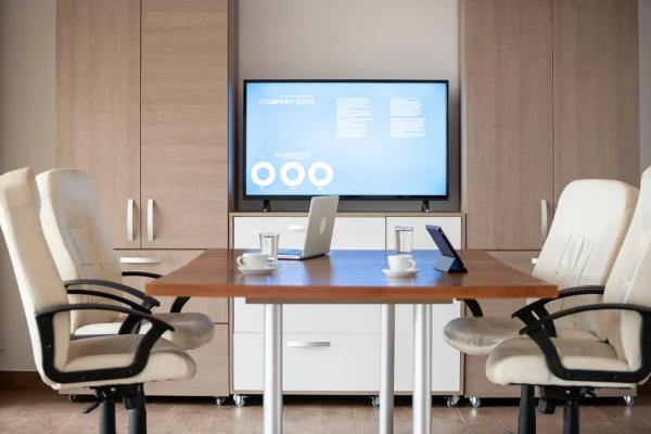 A modern conference room with a large wooden table, four beige office chairs, and a TV displaying "Company Data" along with some charts and text. On the table, there are two cups of coffee, two glasses of water, a laptop, and a tablet, creating a professional setting for meetings or presentations.