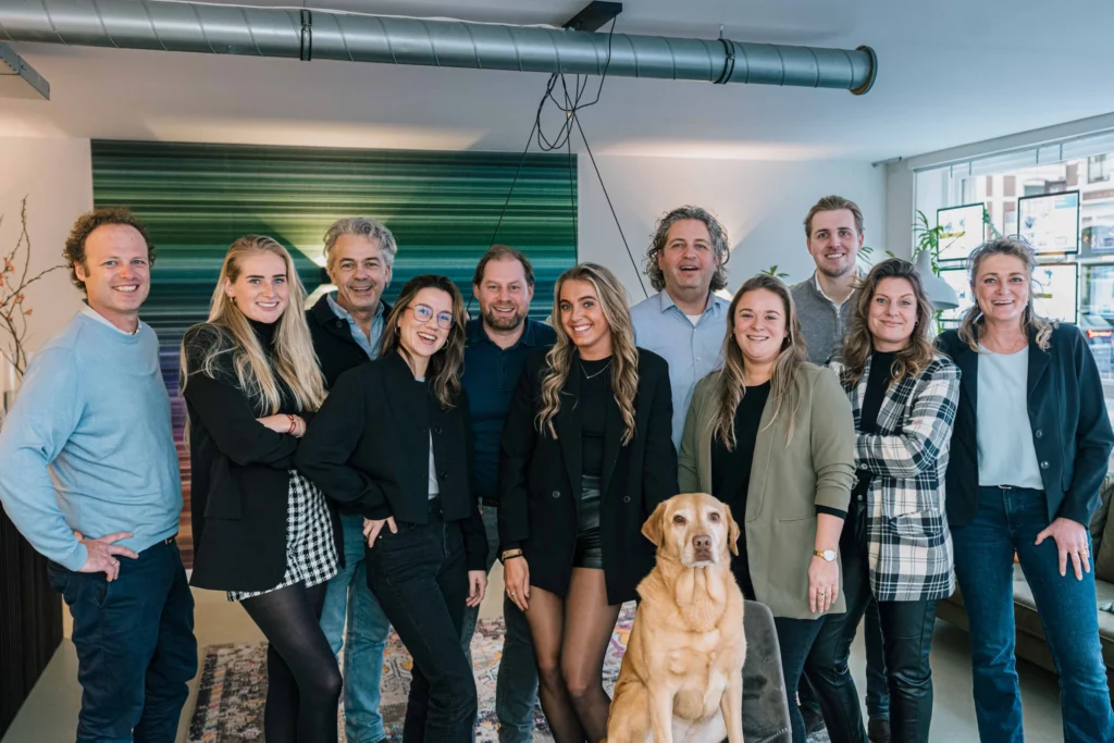 Een groep van elf mensen en een hond poseren samen in een kantoor, allemaal glimlachend naar de camera.