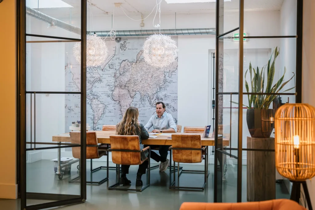 Een man en een vrouw zitten tegenover elkaar aan een grote houten tafel in een modern kantoor met een wereldkaart aan de muur.