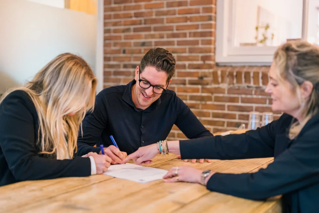 Drie mensen zitten aan een houten tafel, lachen en vullen samen documenten in.