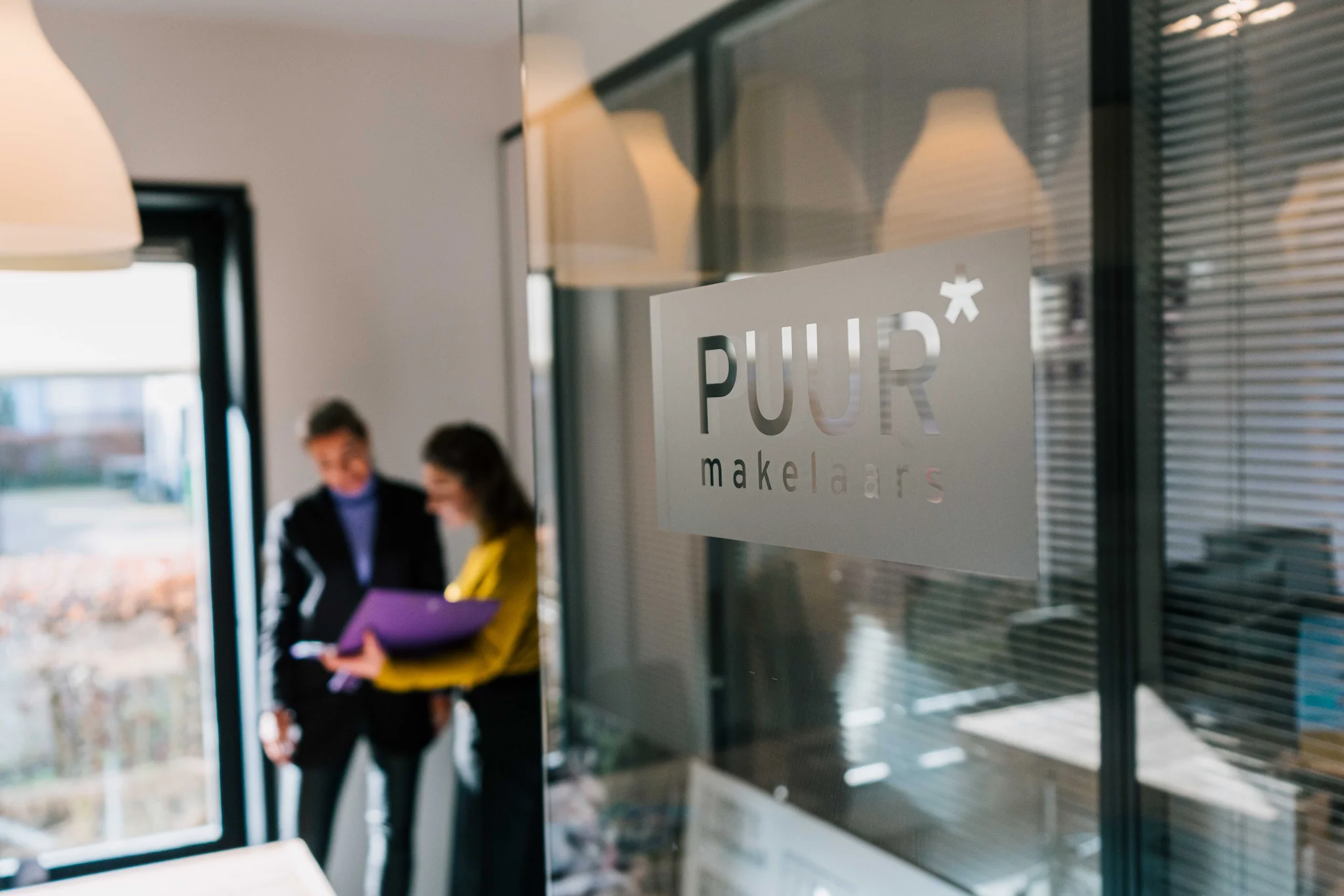 Een matglazen deur met het logo "PUUR makelaars", met twee vrouwen die op de achtergrond iets bespreken.