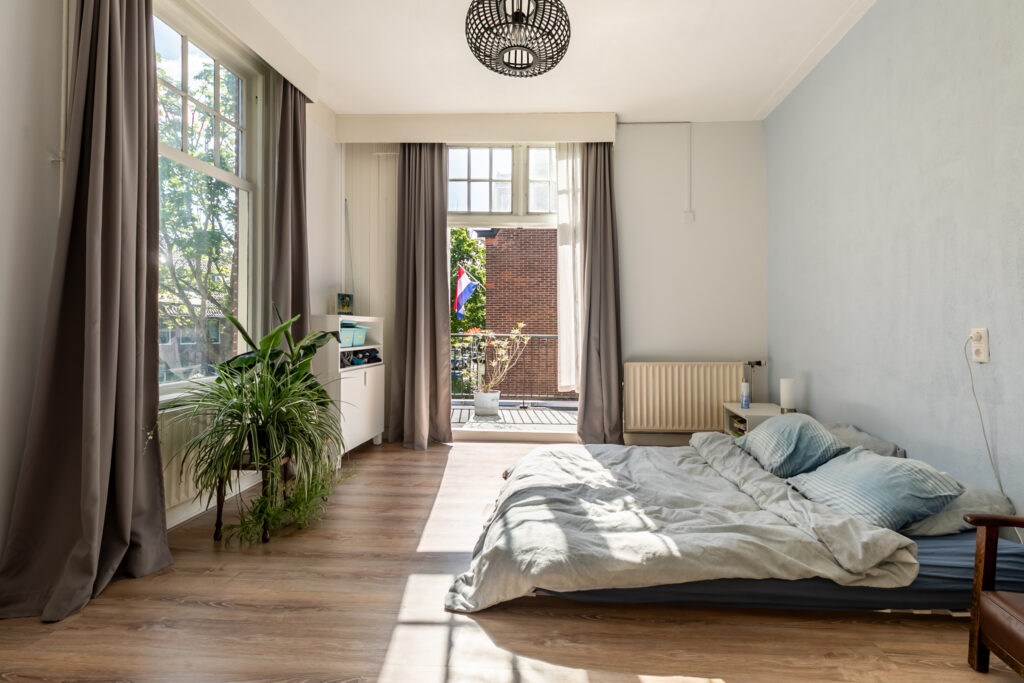 Bright bedroom with bed and plants, open balcony doors.