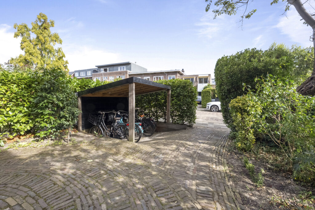Bicycles parked under wooden shelter surrounded by greenery.