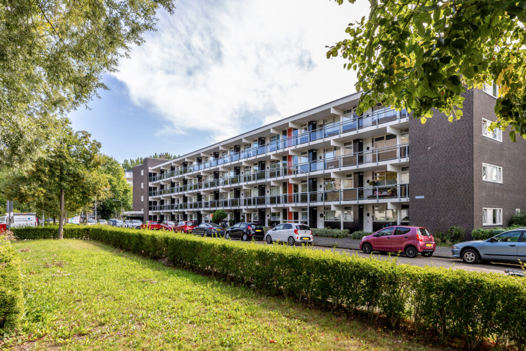 Four-story apartment building with parked cars and greenery.