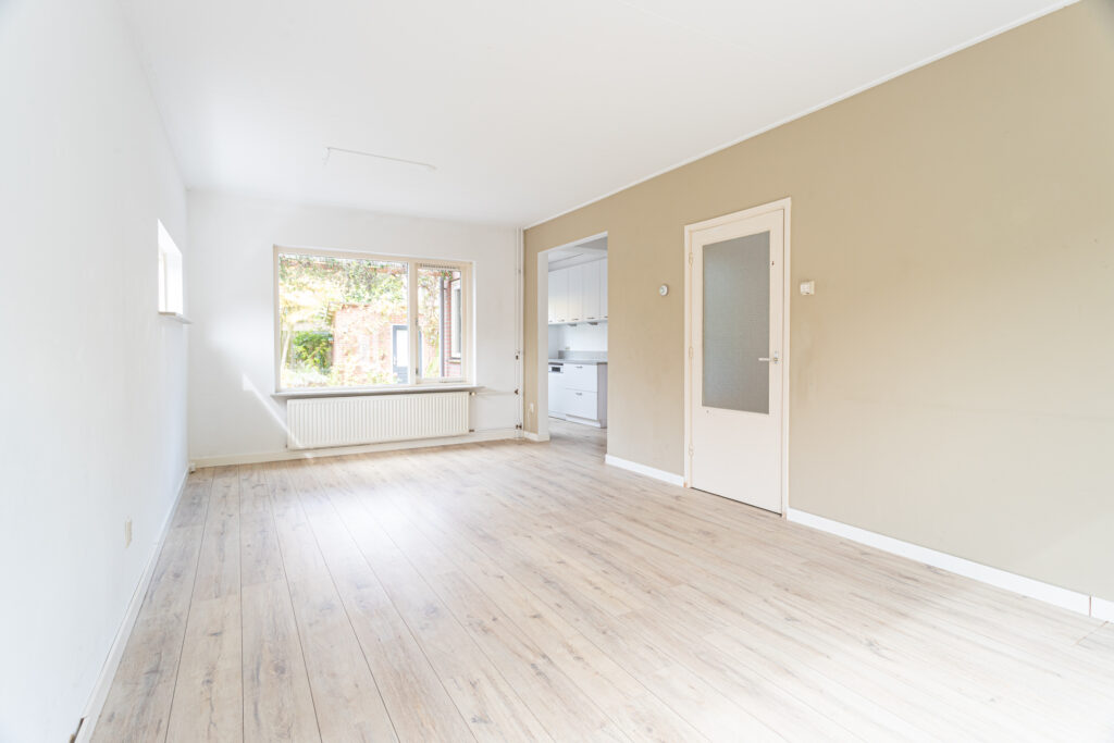 Empty room with wooden floor, beige wall, large window.