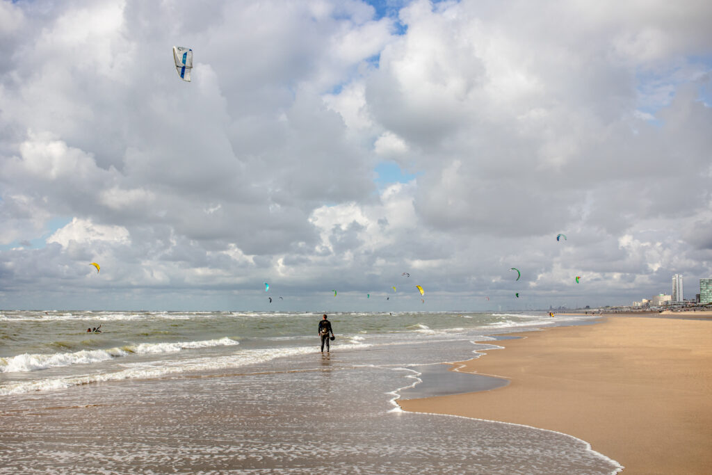 Person walking on beach with kite surfers in background.