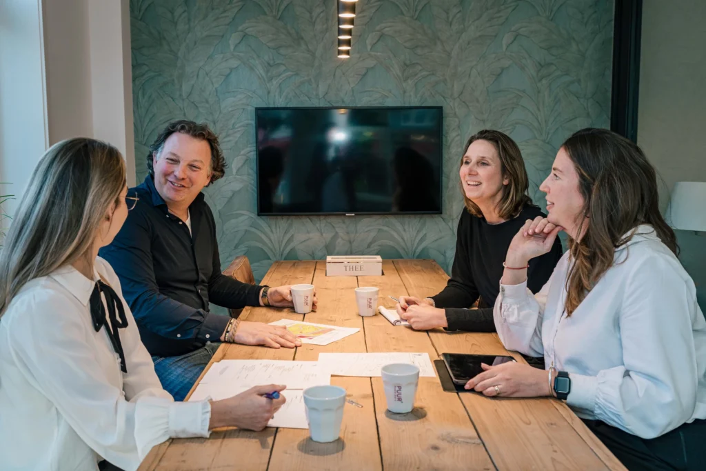 Vier mensen hebben een vergadering rond een houten tafel in een kantoor, glimlachend en pratend met documenten en koffiekoppen voor hen.