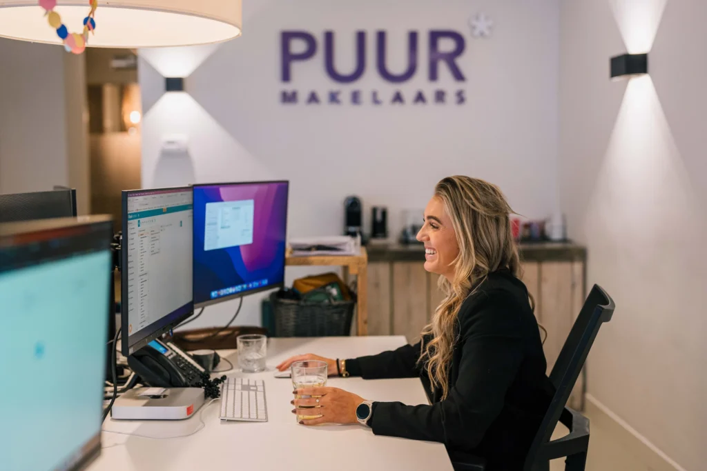 Een vrouw die aan een bureau zit voor meerdere computerschermen, glimlachend en een glas vasthoudend, met het bord "PUUR makelaars" op de muur achter haar.