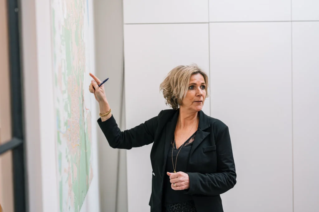 Een vrouw in een zwarte blazer wijst met een pen naar een kaart op de muur.