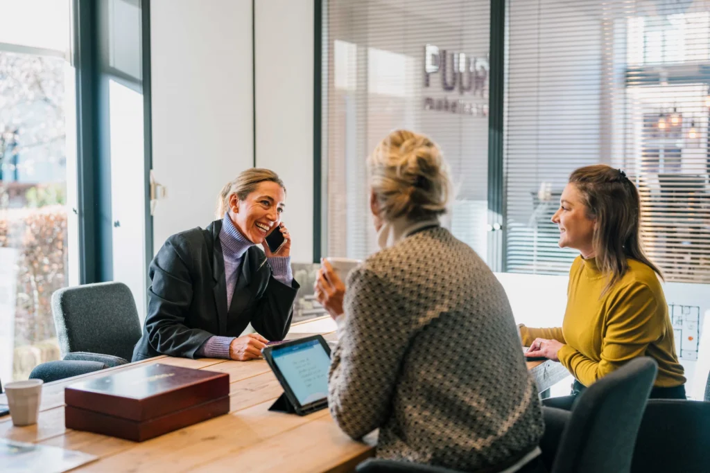 Drie vrouwen hebben een vergadering in een kantoor, een vrouw is aan het telefoneren terwijl de andere twee glimlachen en luisteren.